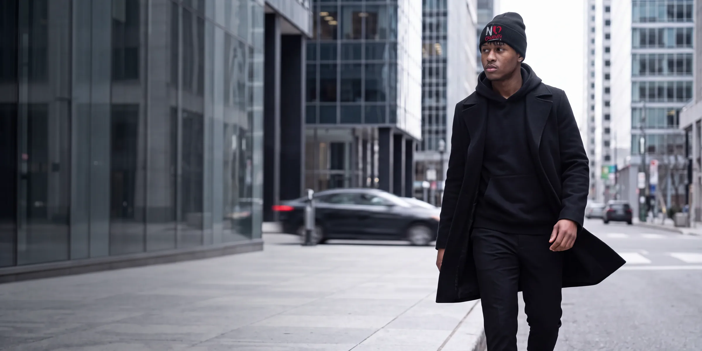 NO CREATIVITY DROP 01: model in the NO CREATIVITY beanie, black hoodie and overcoat walking a downtown curb